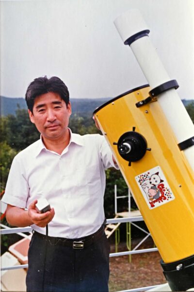 a man in a white shirt and black pants stands next to a yellow telescope