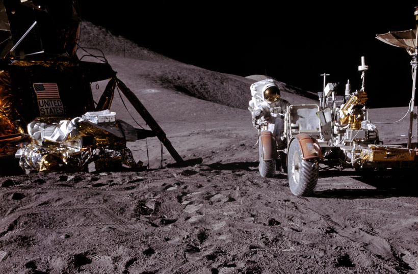 The lander to the left of the image with the rover to the right. An astronaut looks toward the camera from behind the rover.