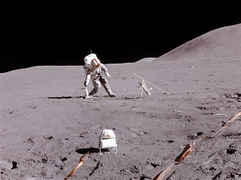 An astronaut on the lunar surface faces the camera and leans to the side to pick up a tool.