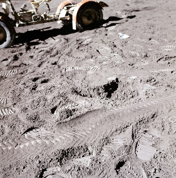 With the rover in the background, the lunar soil is shown with many boot prints and rover tracks. The rover tracks appear to be less deep.