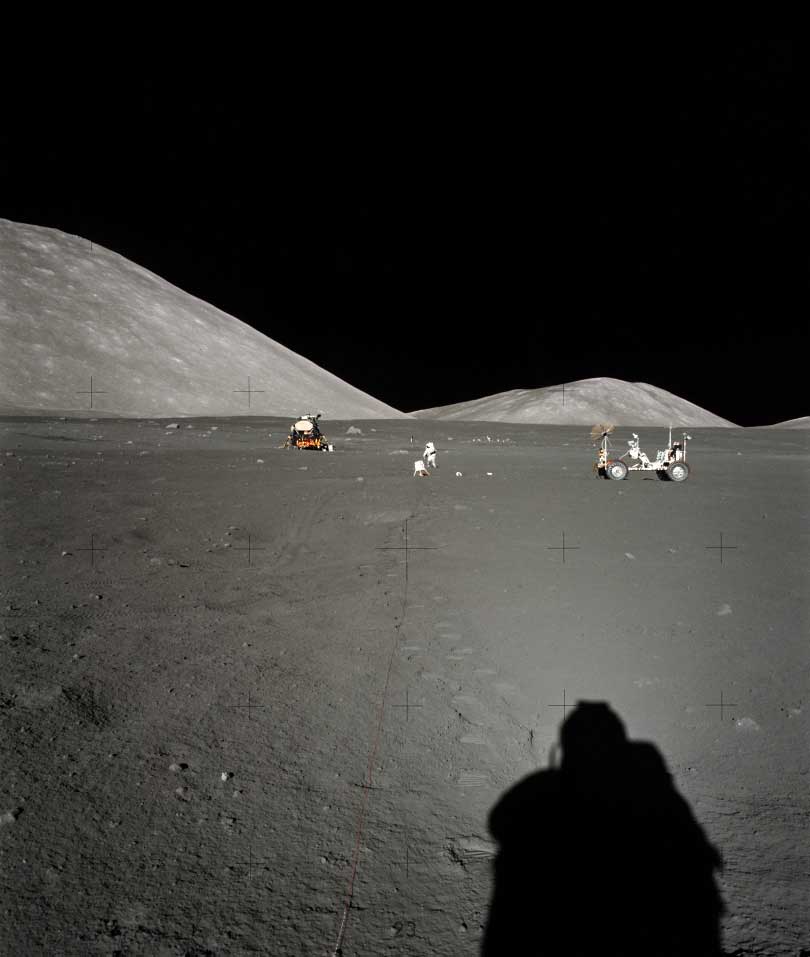 the shadow of a person in the foreground with lunar vehicles far away in front of hills on the moon