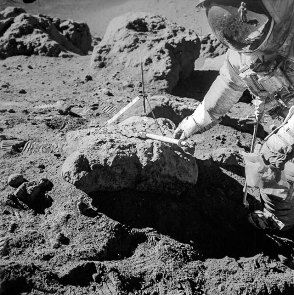 One astronaut leans over a rock and tools while another is reflected in his visor.