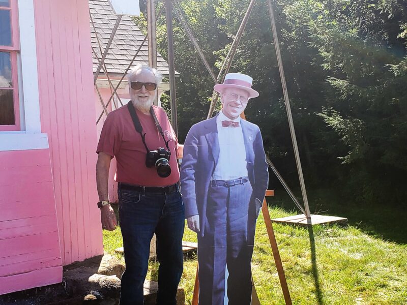 An elderly man standing next to a standee of Russell Porter in front of a pink building and a radio tower