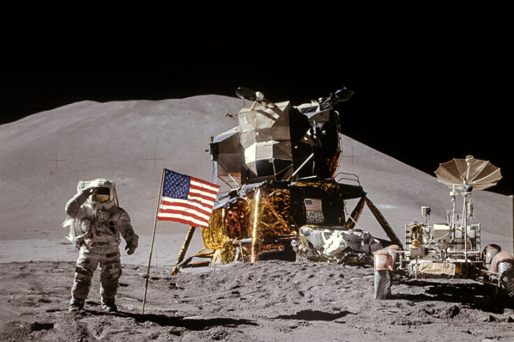 Astronaut stands to the left of American flag on the lunar surface, with hills in the background