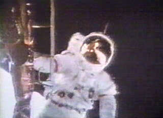 Astronaut in suit facing the camera right near the lander, which is shown in the reflection of his helmet