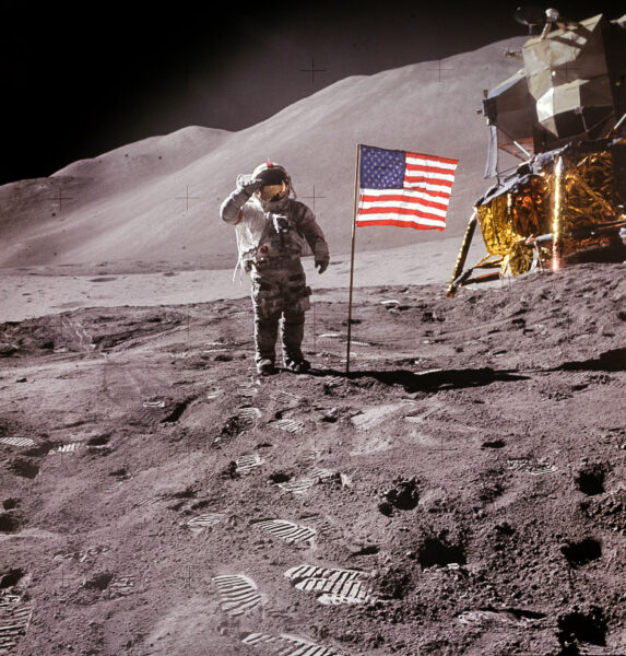 Astronaut stands to the left of American flag on the lunar surface, with hills in the background
