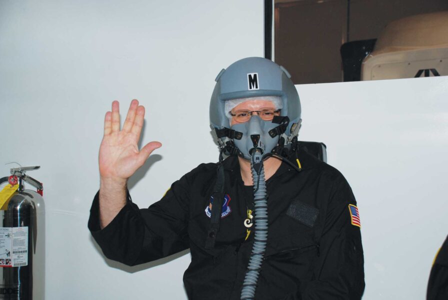 a person with a helmet, face mask, and nozzle waves to the camera in front of a wall