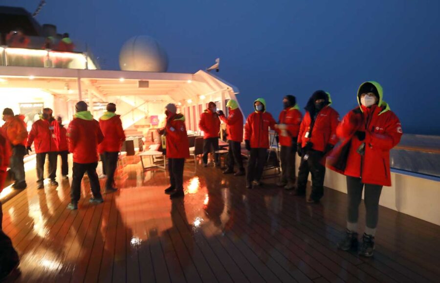a group of people in red jackets stand on a lighted boat deck against a dark blue sky