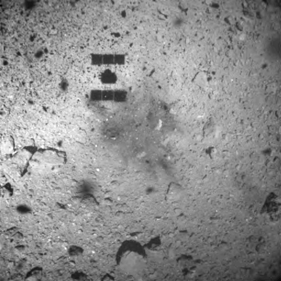 a close up of the asteroid's surface with the faint shadow of a spacecraft over it, in greyscale