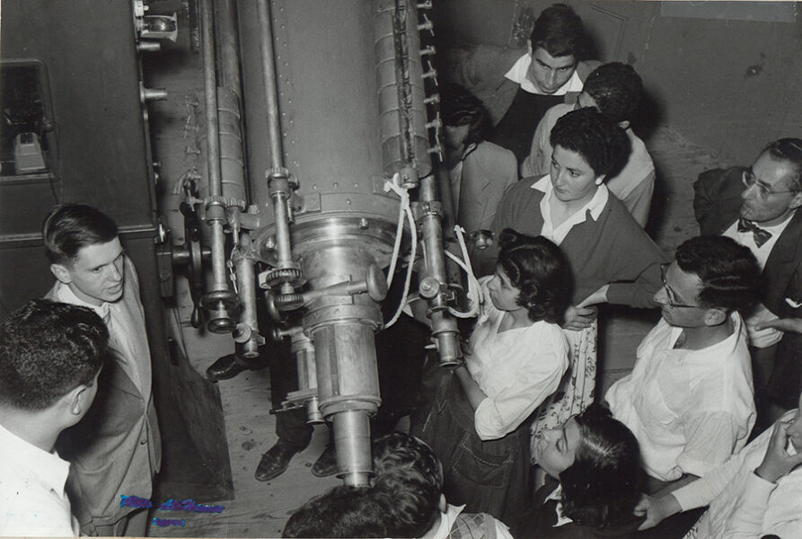 Black and white photo shows group of people surrounding telescope