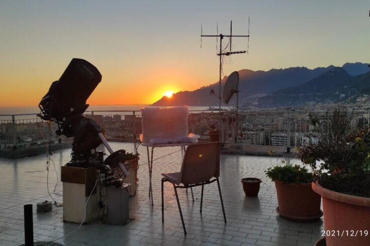 a telescope on a deck oberlooking water with the sun setting behind a mountain
