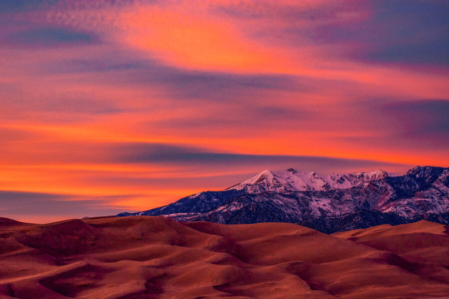 Orange and pink colors of a sunset brighten the sky over the dunes and a snow-capped mountain.