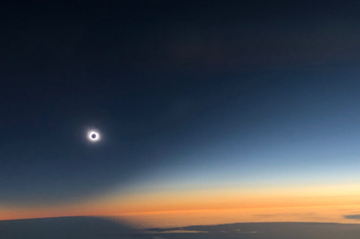 a solar eclipse above a cloud bank with a dark blue sky turning orange towards the horizon