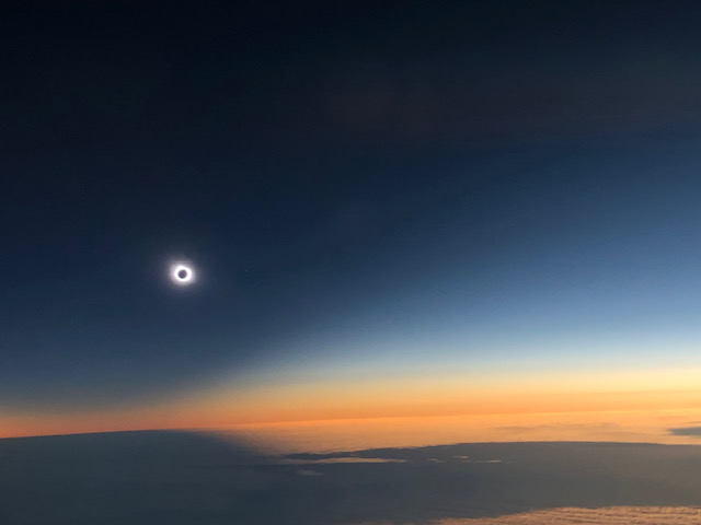 a solar eclipse above a cloud bank with a dark blue sky turning orange towards the horizon